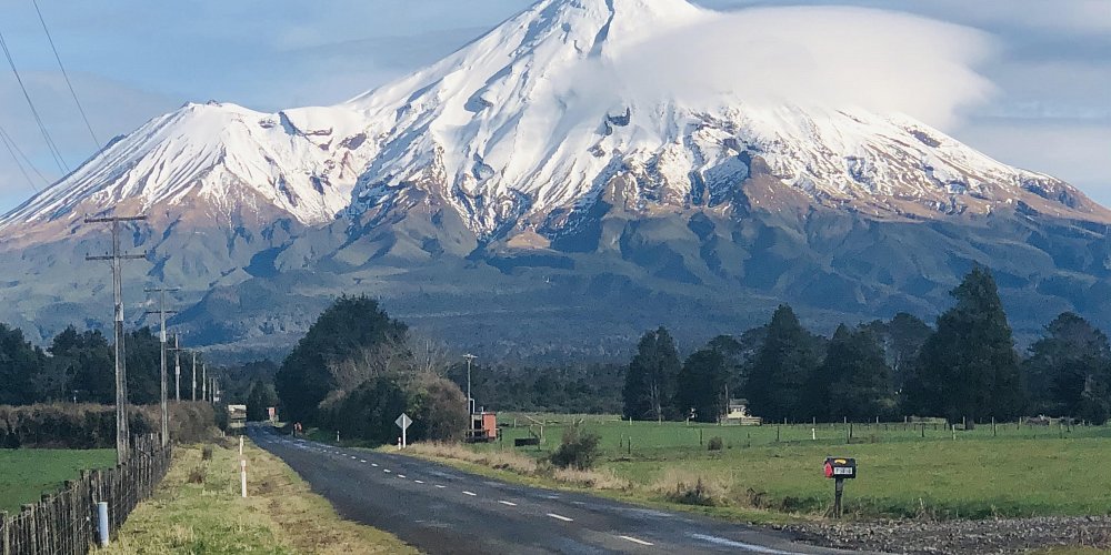 Snow-covered Mount Taranaki rising above farmland with a country road leading toward it, a highlight of the Auckland to Wellington drive via New Plymouth.