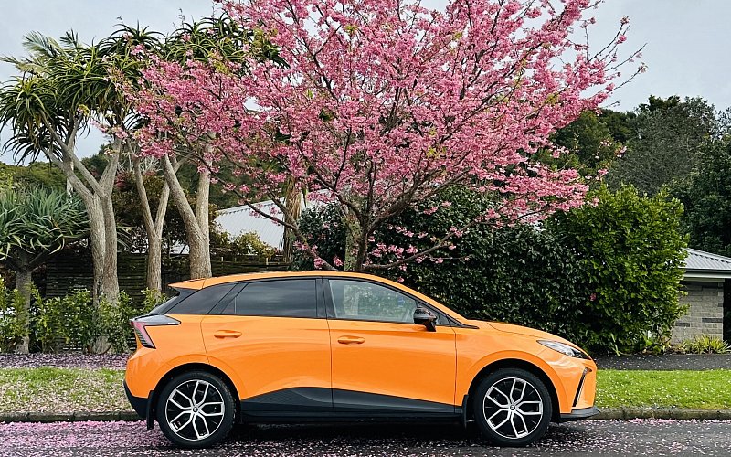 Orange MG4 electric car parked under a cherry blossom tree with pink petals on a suburban street