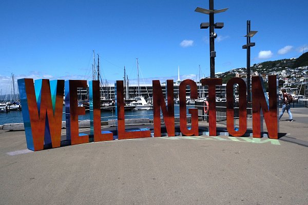 Large Wellington sign on the waterfront promenade with marina, boats, and city hills in the background – finishing point of the Auckland to Wellington drive.